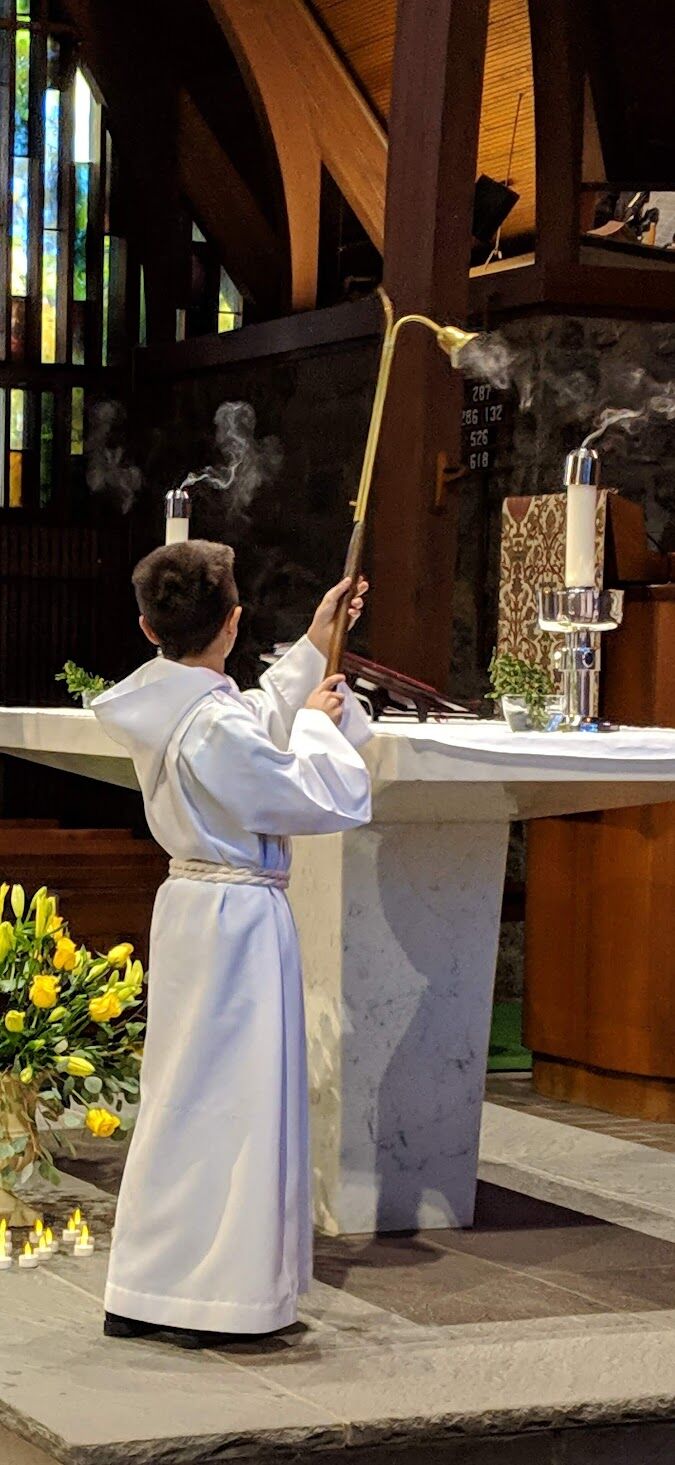 A young acolyte in a white robe reaching up with a brass candle snuffer to extinguish the altar candles