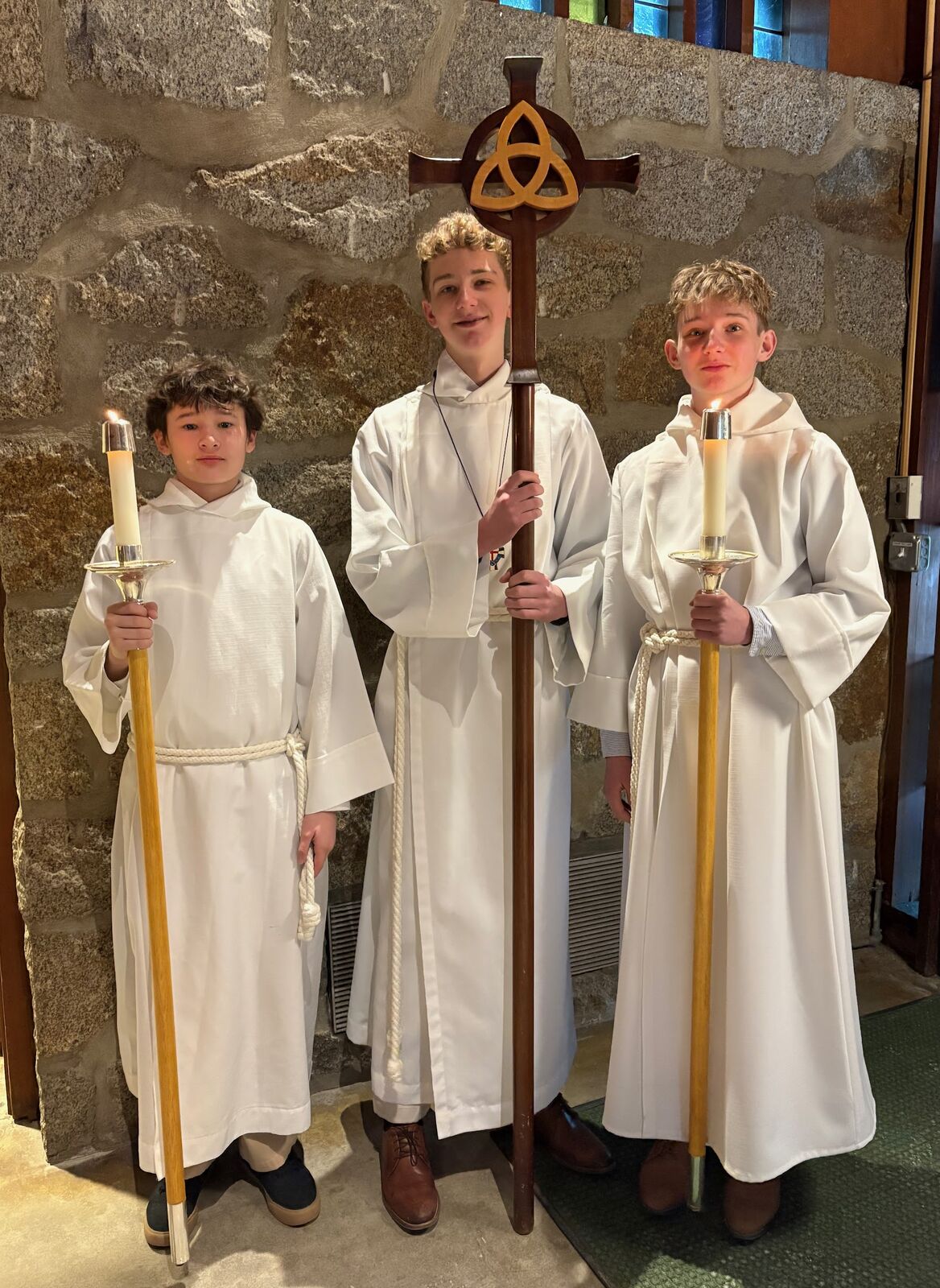 Three young acolytes in white robes holding the processional cross and candles